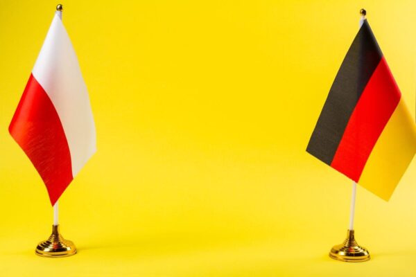 Na żółtym tle stoją obok siebie flagi Polski i Niemiec, symbolizując tłumaczenia z polskiego na niemiecki.