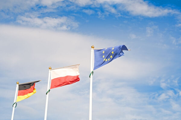 Flagi Niemiec, Polski i Unii Europejskiej powiewają na wietrze, co przypomina o współpracy i potrzebie tłumacza przysięgłego języka niemieckiego.